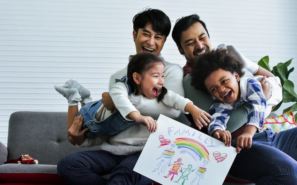Male gay couple taking care of their children who are happy Caucasian girl and African boy, playing with fun, drawing family picture, sitting on sofa in living room at home.