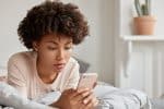 Image of black young lady with Afro haircut checks mobile phone, dressed in casual nightclothes, rests in bed.