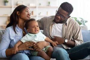Portrait of young African American parents sitting with crying baby son on sofa at home, hugging and calming soothing small baby, man in eyeglasses trying to comfort baby