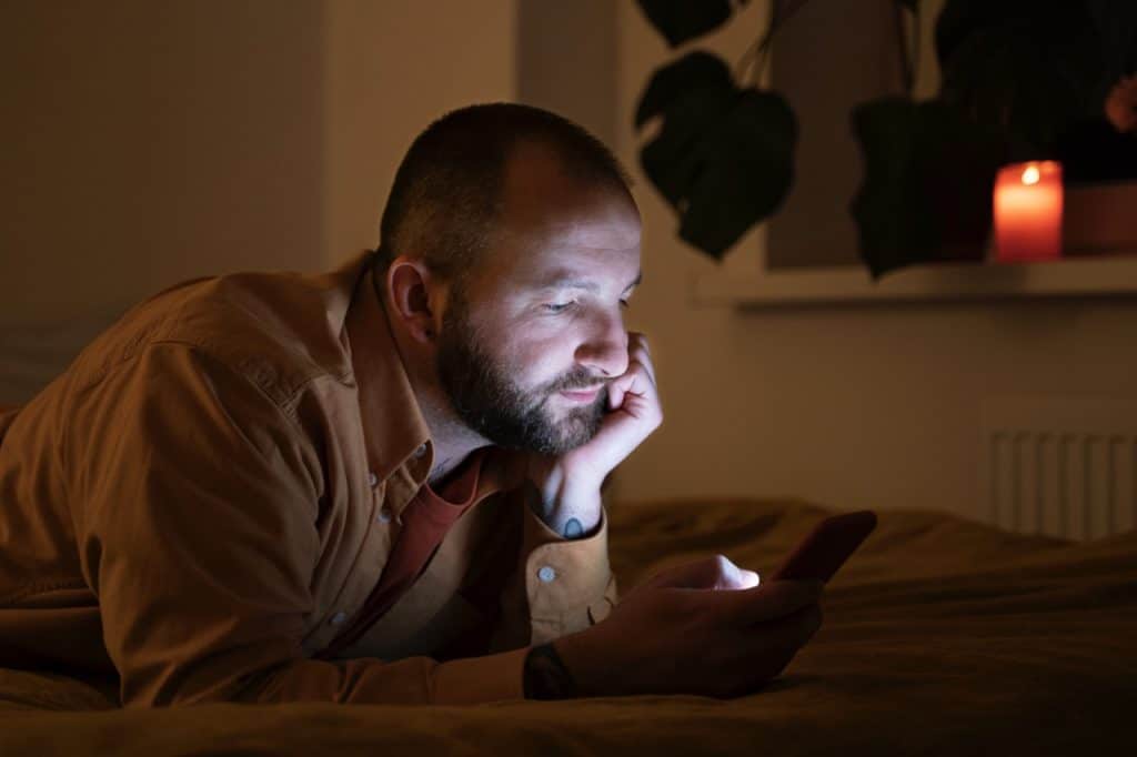 Bald white man with beard lying on bed in dark room looking at smartphone with candle lit. Image by Freepik.