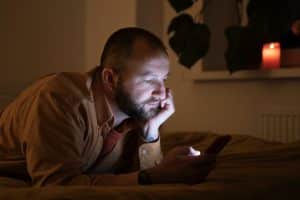 Porn addiction: Bald white man with beard lying on bed in dark room looking at smartphone with lit candle.