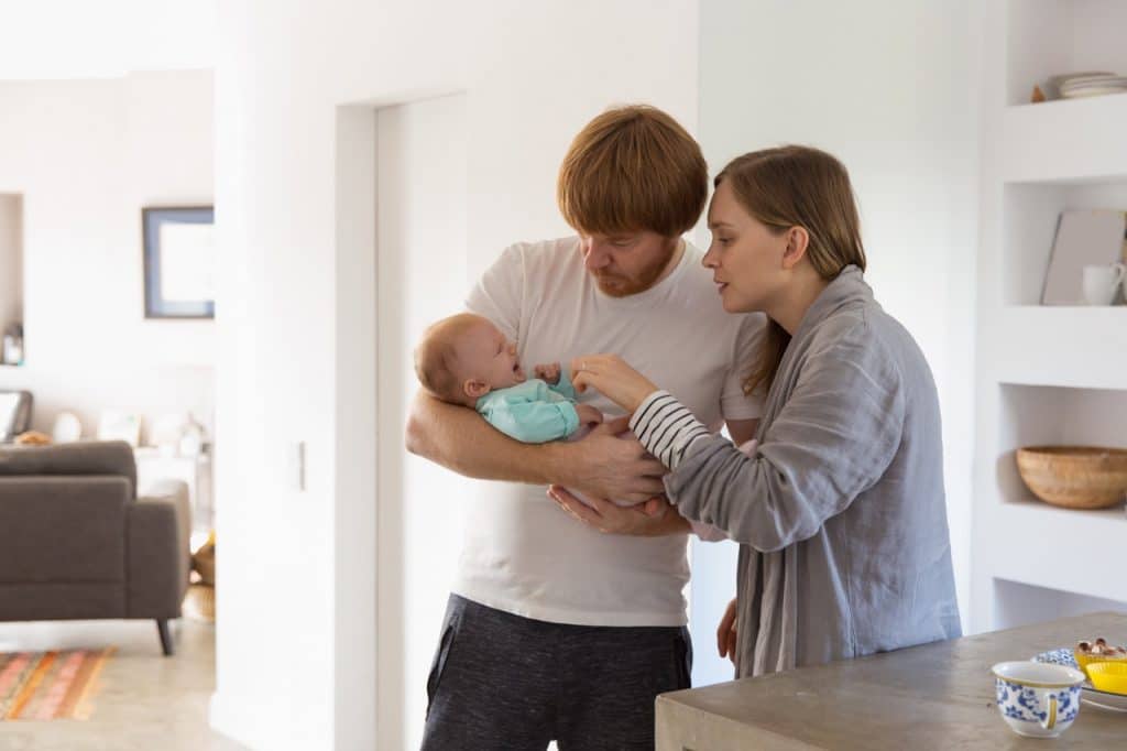 Concerned new parents holding and rocking crying baby. Family portrait of couple and cute little child in home interior.