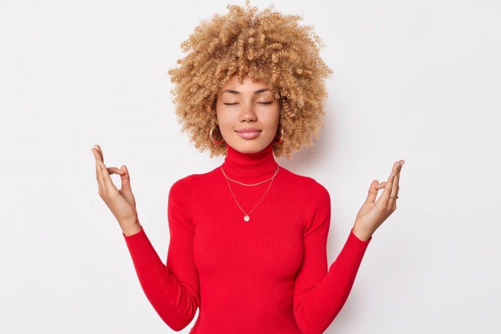 Calm concentrated woman with curly hair keeps eyes closed and meditates indoor makes mudra gesture wears casual red turtleneck isolated over white background tries to relief stress. Zen pose